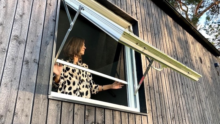 Sieviete raibā kreklā atver lielu uz āru vērstu logu ar RIPO insektu sieti uz koka ēkas sāniem.